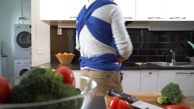 Father with little baby sleeping in sling cooking in the kitchen. Man on paternity leave working household chores. Dad enjoying fatherhood responsibility caring for child