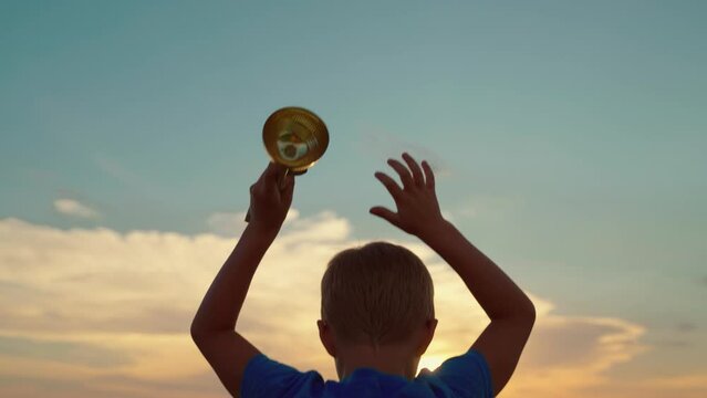 Kid celebrating victory, winner in sports, concept of life success. Slow motion. Winner in business. Silhouette of happy champion child, sporty little boy holding gold cup in his hands against sky.