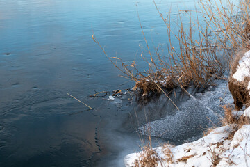 Winter view on cold snowy frozen river shore with dry grass. Water flow on lake covered in snow close-up