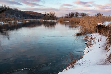 Fototapeta premium Winter view on cold river with scenic colorful clouds with reflection in mirror water and dry reed grass on shore. Zmiyevsky region on Siverskyi Donets River covered in snow in Ukraine