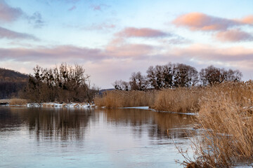 Winter view on cold river with scenic colorful clouds with reflection in mirror water surface and dry reed grass. Zmiyevsky region on Siverskyi Donets River covered in snow in Ukraine