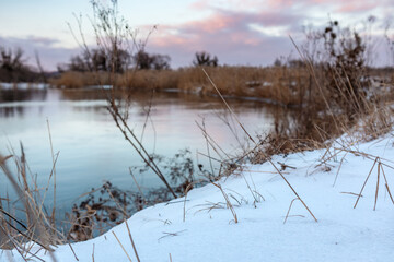 Cold river in winter, snowy close-up with scenic colorful clouds with reflection in water and dry reed grass. Zmiyevsky region on Siverskyi Donets River in Ukraine