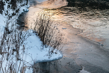 Winter view on cold snowy frozen river with dry grass and sun reflection. Water flow on lake...