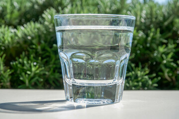Glass of water on white table. Transparent faceted glass on green natural background