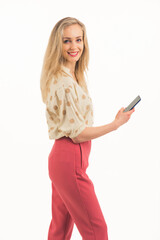 Naklejka premium cute Caucasian blue-eyed blonde girl with a pretty smile wearing a shirt and pink trousers standing sideways and holding a phone studio shot white background medium full shot copy space. High quality