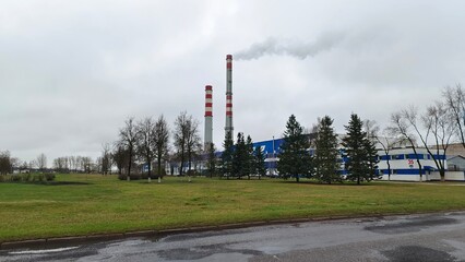 At the edge of town near the paved road is a factory. Smoke is coming out of the factory's pipes. In front of the factory there is a grassy lawn with bushes and trees growing on it. Cloudy