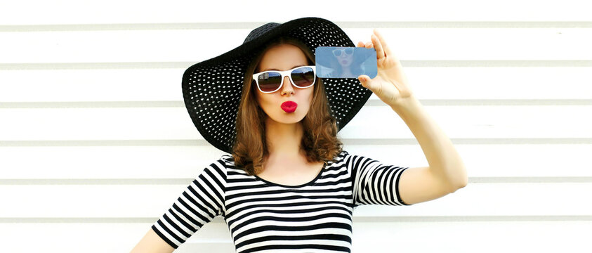 Portrait of beautiful young woman taking selfie by smartphone blowing her lips with red lipstick sending sweet air kiss wearing summer straw round hat, striped shirt on white background