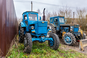 two old blue tractors under an autumn gray sky near a brown fence