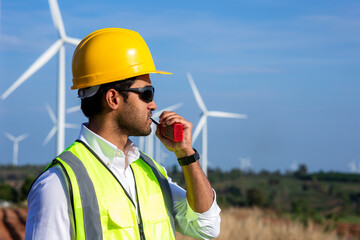 Young engineer wear protective helmet using radio communication in wind turbine farm on background.