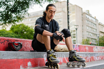 young latin man sitting with rollers on at a skate park buckling protective wrist gloves