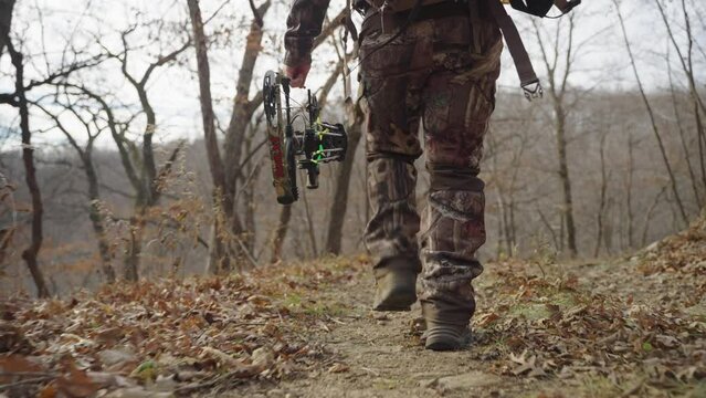 Low angle, bowhunter's legs wearing camouflage clothing holding bow and arrow. Walking on winter forest path full of dead leaves