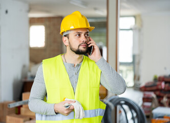 Confident young bearded civil engineer in yellow safety vest and hard hat standing at construction site indoors, solving work issues on phone