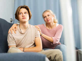 Obraz premium Friendly caring woman calming sad teenage son after disagreements, while sitting together in cozy living room..