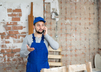 Focused young bearded contractor in blue overall standing at construction site indoors, discussing...