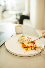 cutlery in hand and a benedict egg on a stylish white plate. a snack in a stylish white cafe. vertical, food content, selective focus