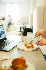 cutlery in hand and a Benedict egg on a stylish white plate and a laptop on the background. working snack in a stylish white cafe, coworking. vertically, food content, selective focus