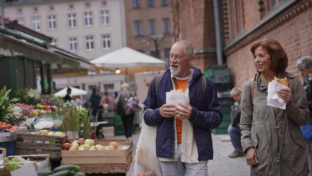 Happy senior couple tourists with snack in town on outdoor market.
