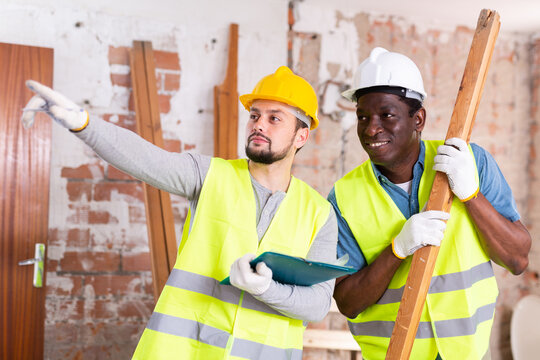 Builders discussing work in cosntruction site indoors. Caucasian man pointing finger, african-american man holding wooden plank.