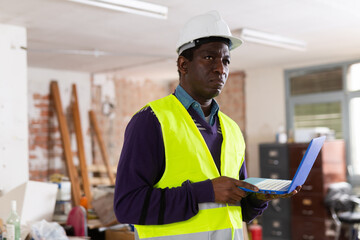 Responsible african american supervisor in yellow safety vest and hard hat inspecting construction site indoors, checking adherence to plans and blueprints on laptop ..
