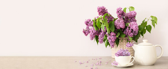 Vase with bouquet of beautiful lilac, teapot and cup on table against light background with space for text