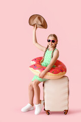 Happy little girl with suitcase, hat and inflatable ring on pink background