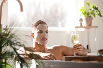 Handsome man with facial mask and glass of wine taking bath at home