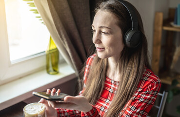 Obraz na płótnie Canvas A woman in a red dress is listening audiobook wearing headphones with phone by the window. The concept of modern technological education and the study of books with joy