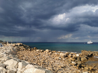 The stone coast of the Mediterranean Sea against the backdrop of the sea with a walking white ship and a dramatic sky.