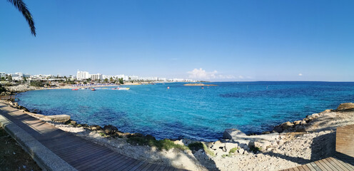 Panoramic view from the rocky coast promenade to the sandy beach of Fig Tree Bay and the island against a blue sky with clouds.