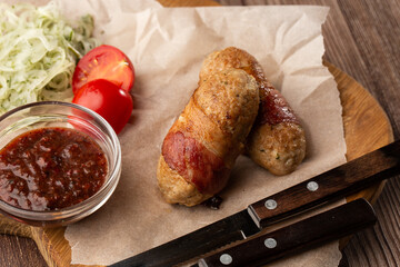 Ready-to-eat pigs sausages wrapped in bacon on wooden board. Fried savory sausages wrapped in bacon served with onion and sauce on wooden background.