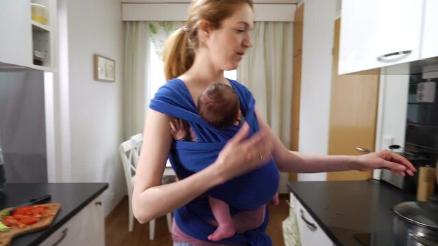 Young mother with baby sleeping in sling cooking in the kitchen. Woman on maternity leave working household chores mom enjoying motherhood responsibility caring for child