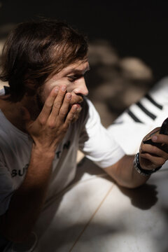 A Young Man Surfer With A Surfboard On The Ocean, Preparing To Surf, Applies Protective Zinc On His Face.