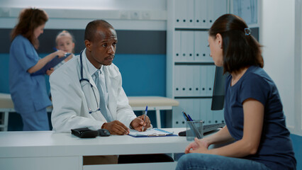 Male doctor having conversation with mother at examination appointment, taking notes on clipboard papers to give prescription medicine as recovery treatment. Medic and parent talking