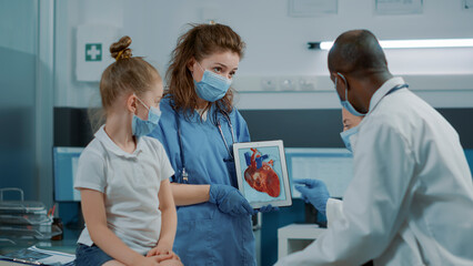 Naklejka na ściany i meble Woman nurse showing cardiology image on digital tablet, letting doctor explain cardiovascular diagnosis to child and parent. Assistant holding modern device with heart anatomy picture.
