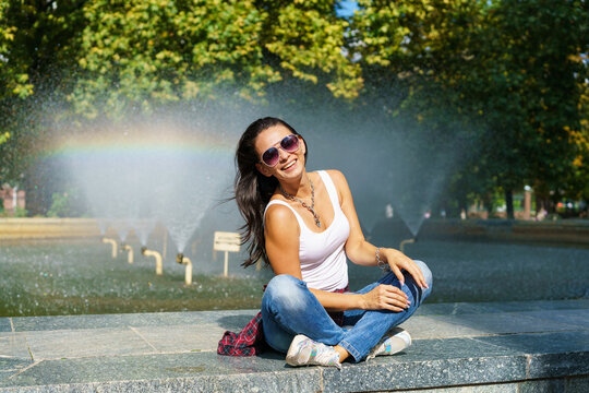 Cheerful young woman in sunglasses sits against backdrop fountain with rainbow and enjoys a sunny day in the park gesturing with her hands in happiness