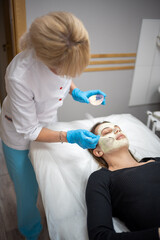 Doctor cosmetologist or dermatologist making face mask in cosmetology salon. Professional Beautician applying face mask on caucasian woman face lying on bed in bathrobe.