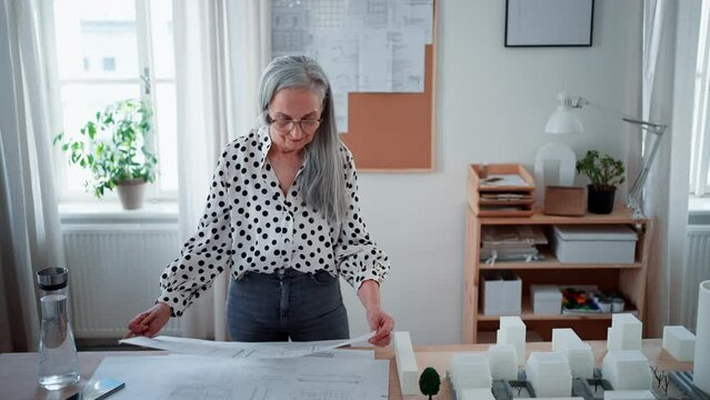 Senior woman architect with model of houses looking at blueprints in office.