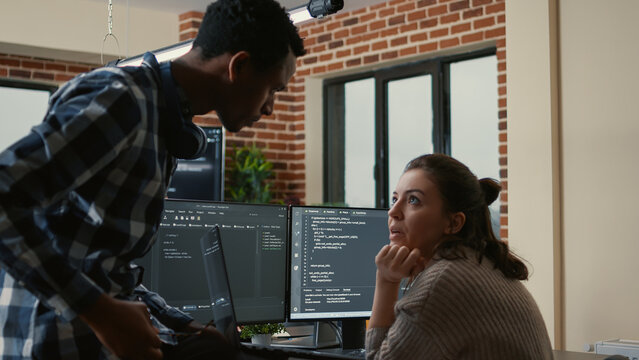 Junior developer sitting down on desk showing laptop with source code to senior dev asking for opinion. Programer writing algorithm interrupted by colleague wanting help with fixing errors.