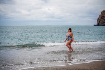 A plump woman in a bathing suit enters the water during the surf. Alone on the beach, Gray sky in the clouds, swimming in winter.