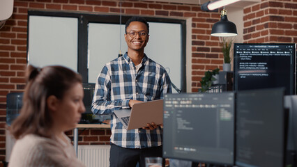 Portrait of african american software developer holding laptop looking up and smiling at camera....