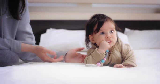 Funny babu girl in a sand bodysuit is lying on her stomach on a white bed in the nursery next to her mom, licking a nipple holder.The concept of love, parenthood, childhood, life, motherhood