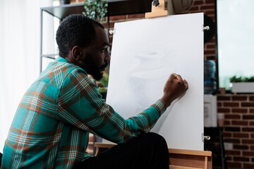 Fototapeta premium African american artist drawing vase model on white canvas working at illustration technique using graphic pencil during art lesson in creativity studio. Man sketching masterprice developing new skill