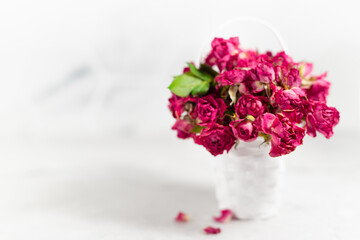 Vintage pink dry tiny roses in white wicker basket in airy soft light white interior with sunbeams, closeup, copy space, blur. Holiday floral background with love, memory and nostalgia.