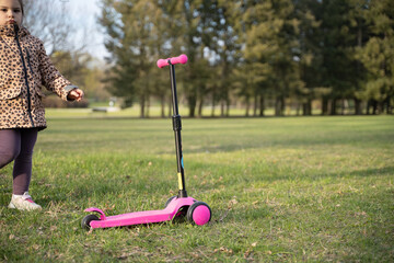 Caucasian preschool girl portrait with pink scooter in the park on sunny cold day in spring. Childhood activity outdoors
