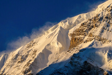 A close up view on snow caped Himalayan peak seen from Annapurna Circuit Trek, Nepal. Sharp and steep slopes of the mountain. Powder snow being blown by strong wind. First sunbeams reaching the peak