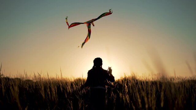 Happy child. Girl with flying kite in park. Child runs at sunset on grass in park. Silhouette of girl with flying kite. Child launches flying kite into sky in the wind. Silhouette of child in meadow