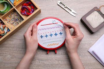 Woman doing different stitches with colorful threads on fabric in embroidery hoop at wooden table,...