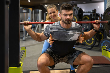 Fototapeta na wymiar Muscular young sporty man lifting heavy weights with assistance of attractive blonde fit woman at gym
