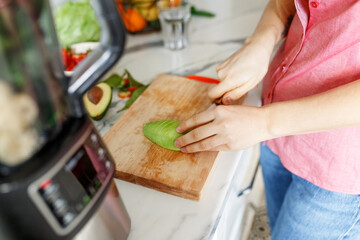 Portrait of beautiful woman cutting avocado for smoothies with blender. Healthy eating, dieting lifestyle concept,preparing drink with bananas, avocado and spinach at home in kitchen, mixer on table.