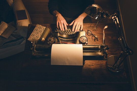 Typing hands of a writer looking for inspiration to start a new novel on his vintage typewriter in a retro style studio: a wooden desk lit by a lamp, old books and a pair of glasses.